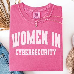 May include: A pink short-sleeve t-shirt with the white text "WOMEN IN CYBERSECURITY" printed in a collegiate font across the chest. The shirt features a Comfort Colors brand label at the inner neckline and a crew neck design.