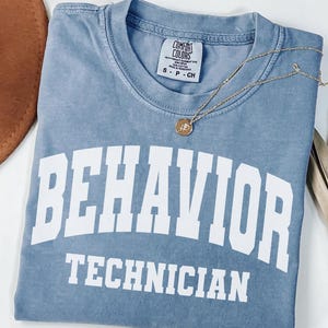 May include: A blue jean colored short sleeve t-shirt with a crew neckline. The shirt features the white text BEHAVIOR TECHNICIAN printed in a bold, arched collegiate font across the chest. A Comfort Colors brand label is visible at the inner neck.