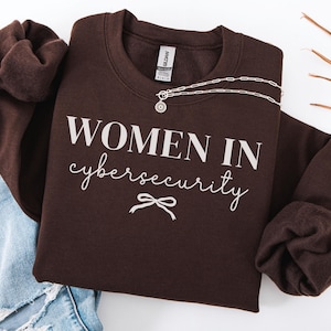 May include: A dark chocolate brown crewneck sweatshirt featuring the white text "WOMEN IN cybersecurity" printed on the front, accompanied by a small white bow graphic below the text. A silver-toned chain necklace rests on the collar area.
