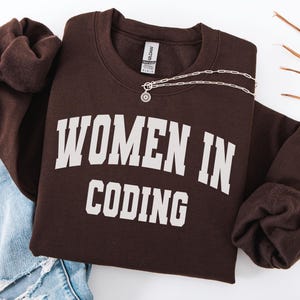 May include: A dark chocolate brown long-sleeve sweatshirt featuring the text "WOMEN IN CODING" in a large, white, arched collegiate-style font across the chest. The garment has a crew neckline and a standard fit, displayed with a silver chain necklace.