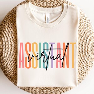 May include: Cream-colored t-shirt with the words "ASSISTANT virtual" in colorful block letters and black script. The shirt is displayed on a woven, circular surface.
