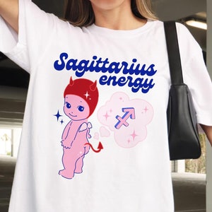 Puede incluir: Camiseta blanca con un diablo rosa de dibujos animados con cuernos rojos y cola. El diablo sostiene un coraz&oacute;n y tiene un globo de pensamiento con el s&iacute;mbolo de Sagitario y el texto "Sagittarius energy".