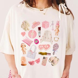 May include: A white t-shirt with a pink and white graphic print of various items, including a heart, a doll, a phone, a pair of slippers, and a pink bow.