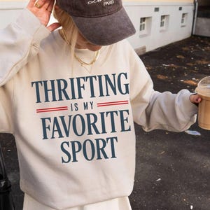 Puede incluir: Una sudadera crema con el texto "THRIFTING IS MY FAVORITE SPORT" en azul marino y rojo. Una gorra de béisbol marrón con "La Paris" bordado en blanco. La persona lleva un collar dorado y sostiene un café.