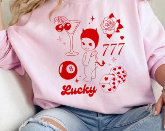 May include: Pink sweatshirt with a red graphic design featuring a devil, a rose, a cocktail glass, cherries, a pool ball, dice, and the text "777" and "Lucky".