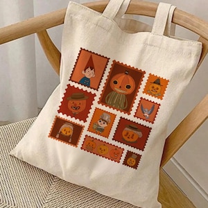 Könnte beinhalten: Beige Canvas-Tragetasche mit Halloween-Design. Das Design zeigt verschiedene orangefarbene und braune Illustrationen im Postkartenstil von Kürbissen, Figuren und anderen gruseligen Elementen. Die Tasche hat zwei Griffe und liegt auf einem Holzstuhl.