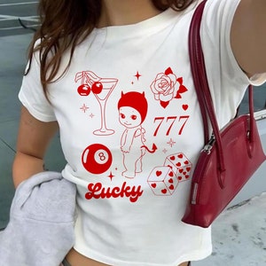 Puede incluir: Camiseta blanca con un diseño gráfico rojo que presenta un bebé diablo, una cereza, una copa de martini, una rosa, una bola de billar, dados y las palabras "777" y "Lucky".