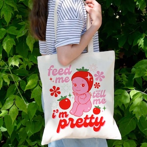 May include: A white tote bag with a pink and red cartoon strawberry character. The text on the bag reads "feed me and tell me i'm pretty".