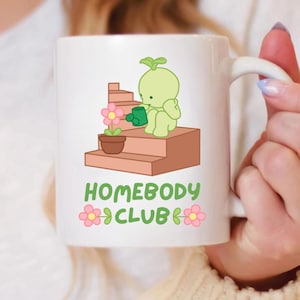 May include: White ceramic mug featuring a cartoon illustration of a green sprout watering a flower on a set of stairs. The mug has the text "HOMEBODY CLUB" with pink flower accents.