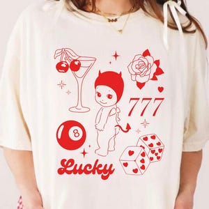 May include: A white oversized t-shirt with a red graphic design featuring a cartoon devil holding a martini glass with cherries, a rose, a dice, a pool ball, and the word "Lucky".