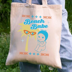 May include: Beige tote bag with the words "Beach Babe" in blue script. The bag features a cartoon palm tree with sunglasses and a baby in a blue swim cap. Decorative patterns in orange and blue complete the design.
