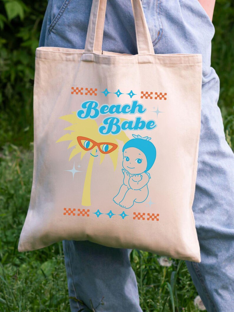 May include: Beige tote bag with the words "Beach Babe" in blue script. The bag features a cartoon palm tree with sunglasses and a baby in a blue swim cap. Decorative patterns in orange and blue complete the design.