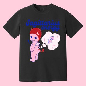 Puede incluir: Camiseta negra con un dibujo animado de diablo rosa con cuernos rojos y un sombrero rojo. El diablo tiene una burbuja de pensamiento sobre su cabeza con un s&iacute;mbolo del zod&iacute;aco Sagitario azul. El texto "Sagittarius energy" est&aacute; impreso en azul en la camiseta.