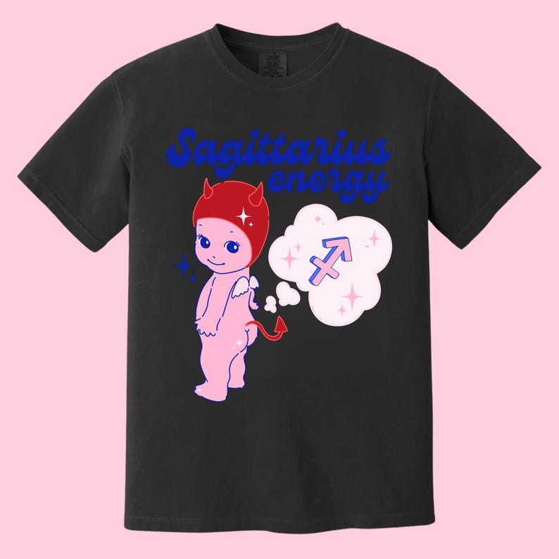 Puede incluir: Camiseta negra con un dibujo animado de diablo rosa con cuernos rojos y un sombrero rojo. El diablo tiene una burbuja de pensamiento sobre su cabeza con un s&iacute;mbolo del zod&iacute;aco Sagitario azul. El texto "Sagittarius energy" est&aacute; impreso en azul en la camiseta.