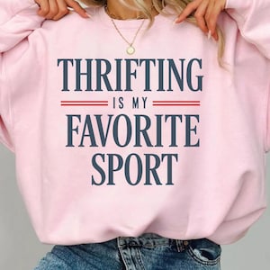 May include: A light pink sweatshirt with the text "THRIFTING IS MY FAVORITE SPORT" in navy blue, with red lines separating the phrases. The sweatshirt is a casual style, suitable for everyday wear.