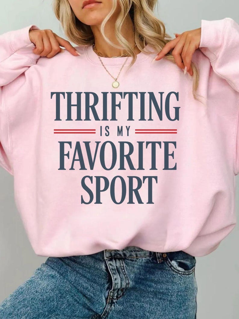 May include: A light pink sweatshirt with the text "THRIFTING IS MY FAVORITE SPORT" in navy blue, with red lines separating the phrases. The sweatshirt is a casual style, suitable for everyday wear.