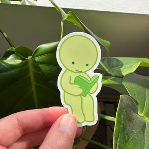May include: A green sticker featuring a cartoon character sitting and reading a book. The character has a round head and a simple body. The book is green and has a white spine.
