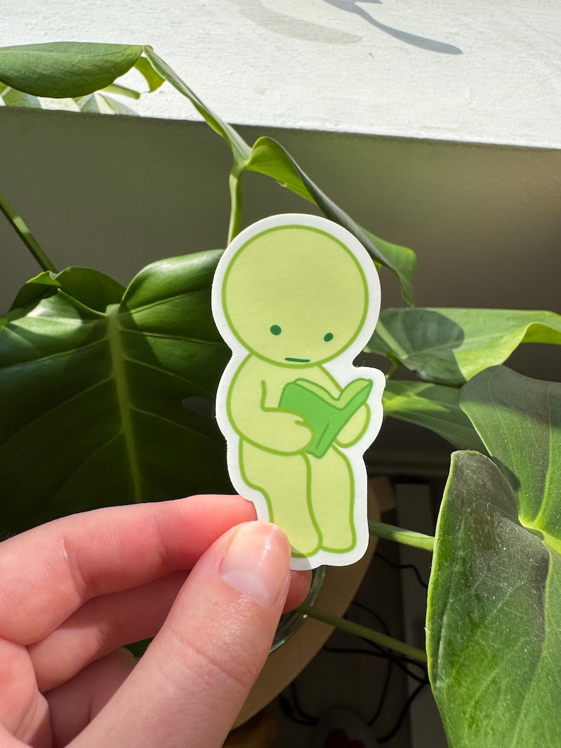 May include: A green sticker featuring a cartoon character sitting and reading a book. The character has a round head and a simple body. The book is green and has a white spine.