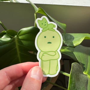May include: A green sticker featuring a cartoon character with a green cat perched on its head. The character has its arms crossed and a worried expression.
