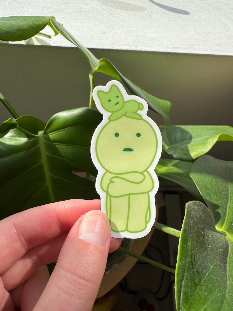 May include: A green sticker featuring a cartoon character with a green cat perched on its head. The character has its arms crossed and a worried expression.