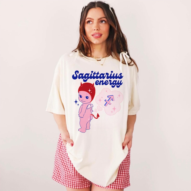 Puede incluir: Camiseta blanca con una ilustraci&oacute;n de dibujos animados de un diablo rosa con cuernos rojos y un halo azul. El texto "Sagittarius energy" est&aacute; impreso en letras azules y rojas. El diablo sostiene una nube rosa con un s&iacute;mbolo del zod&iacute;aco Sagitario azul.