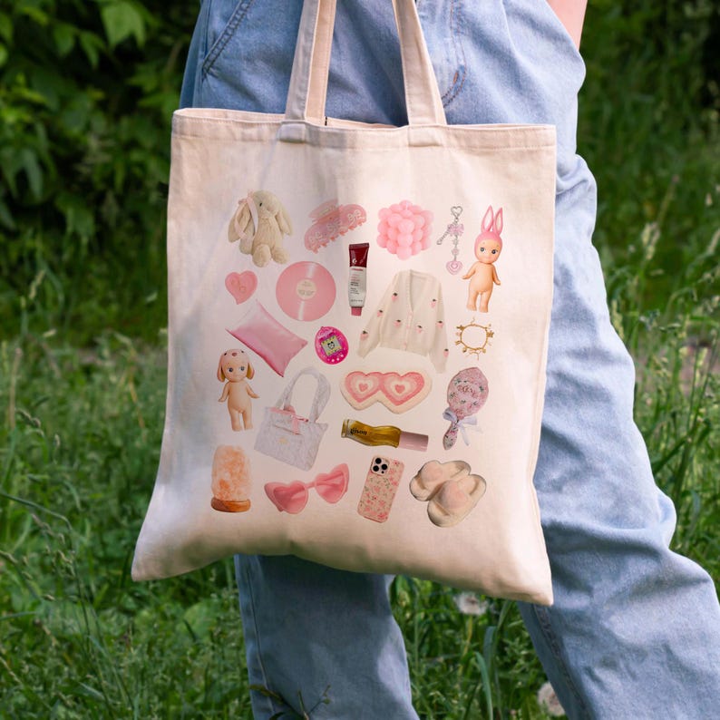 May include: A white canvas tote bag with a pink and white design featuring various items, including a bunny, a phone, a pair of sunglasses, a heart-shaped eye mask, a doll, and a hair clip.