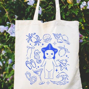 May include: Off-white canvas tote bag featuring a blue illustration of a starfish-headed figure, seashells, a crab, and other beach-themed elements. The bag has two long handles for carrying over the shoulder.
