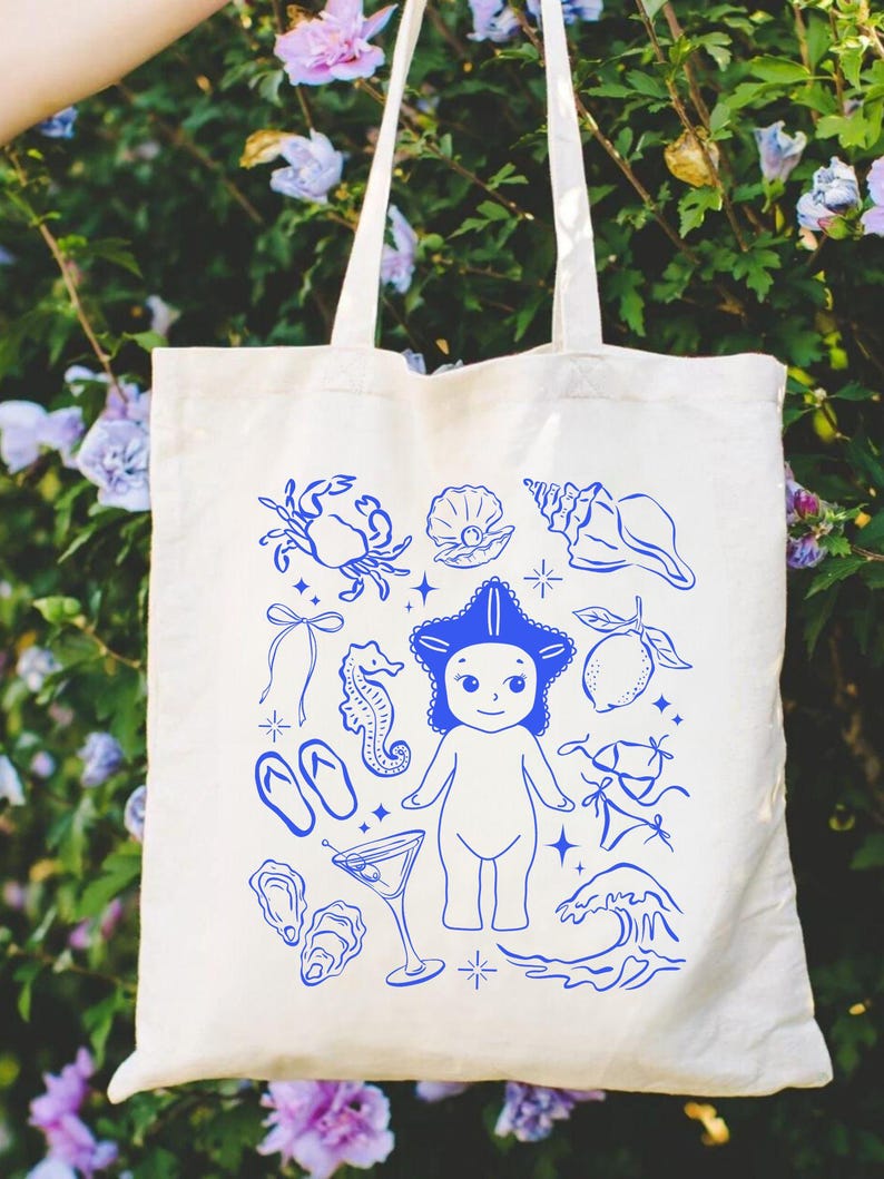 May include: Off-white canvas tote bag featuring a blue illustration of a starfish-headed figure, seashells, a crab, and other beach-themed elements. The bag has two long handles for carrying over the shoulder.