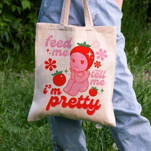 May include: A white tote bag with a pink cartoon character wearing a red tomato hat. The bag has the text "feed me and tell me i'm pretty" in red and pink.