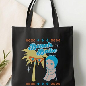 May include: Black tote bag featuring a retro graphic of a palm tree wearing sunglasses and a baby in a blue bonnet. The text "Beach Babe" is in blue script. The design is framed with orange and blue geometric patterns.