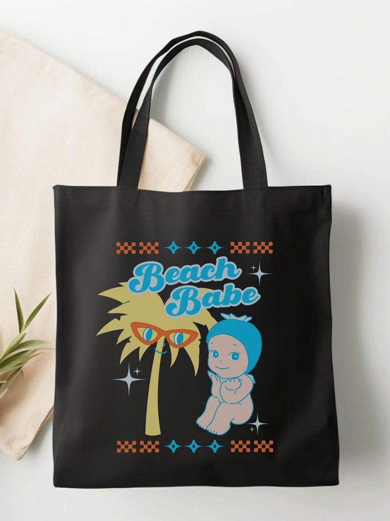 May include: Black tote bag featuring a retro graphic of a palm tree wearing sunglasses and a baby in a blue bonnet. The text "Beach Babe" is in blue script. The design is framed with orange and blue geometric patterns.