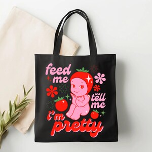 May include: Black canvas tote bag with a pink cartoon tomato character wearing a red tomato hat. The text "feed me and tell me i'm pretty" is printed on the bag.