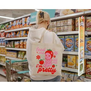 May include: A white tote bag with a pink cartoon character wearing a red tomato hat. The bag has the text "Feed me, tell me I'm pretty" in pink and red letters.