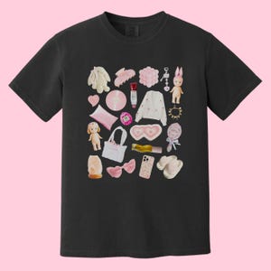 May include: Black t-shirt with a pink and white graphic print of various items, including a doll, a phone, a headband, a makeup brush, a bottle of lotion, a pair of heart-shaped sunglasses, a phone case, a pair of slippers, a small bag, a toy, a ring, a hair clip, a small bag, and a toy bunny.