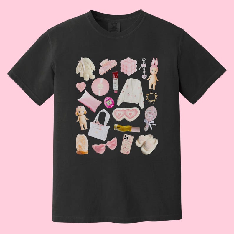 May include: Black t-shirt with a pink and white graphic print of various items, including a doll, a phone, a headband, a makeup brush, a bottle of lotion, a pair of heart-shaped sunglasses, a phone case, a pair of slippers, a small bag, a toy, a ring, a hair clip, a small bag, and a toy bunny.