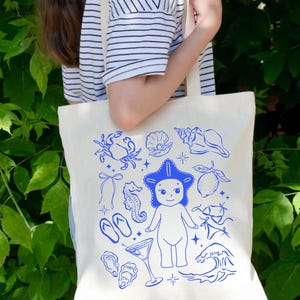 May include: Cream-colored tote bag with a blue illustration of beach-themed elements. The design includes a starfish-headed figure, seashells, a crab, a cocktail, and other nautical images. The bag has a simple, rectangular shape with shoulder straps.