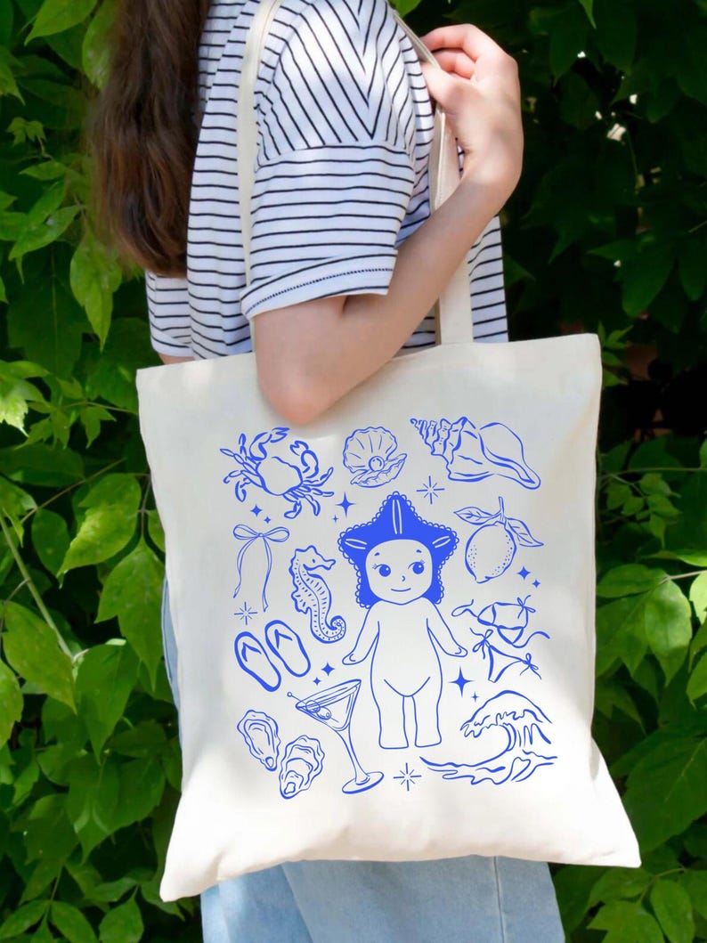 May include: Cream-colored tote bag with a blue illustration of beach-themed elements. The design includes a starfish-headed figure, seashells, a crab, a cocktail, and other nautical images. The bag has a simple, rectangular shape with shoulder straps.