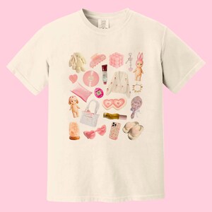 May include: A white t-shirt with a pink and white collage of various items, including a bunny, a hair clip, a phone, a heart-shaped eye mask, a doll, a record, a purse, and a pair of sunglasses.
