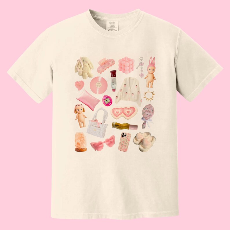 May include: A white t-shirt with a pink and white collage of various items, including a bunny, a hair clip, a phone, a heart-shaped eye mask, a doll, a record, a purse, and a pair of sunglasses.