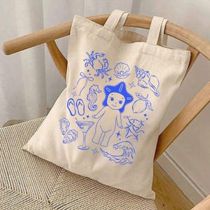May include: A beige canvas tote bag with blue illustrations of sea creatures and beach-themed elements. The design includes a crab, seashell, seahorse, starfish, flip-flops, and a cartoon figure. The bag has two handles and is sitting on a wooden chair.