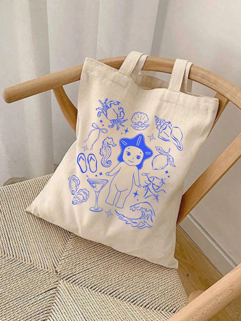 May include: A beige canvas tote bag with blue illustrations of sea creatures and beach-themed elements. The design includes a crab, seashell, seahorse, starfish, flip-flops, and a cartoon figure. The bag has two handles and is sitting on a wooden chair.