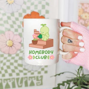 May include: White ceramic mug featuring a cartoon illustration of a green character watering a flower on a set of steps. The mug has the text "HOMEBODY CLUB" in green and pink. The mug is held by a person wearing a pink sweater.