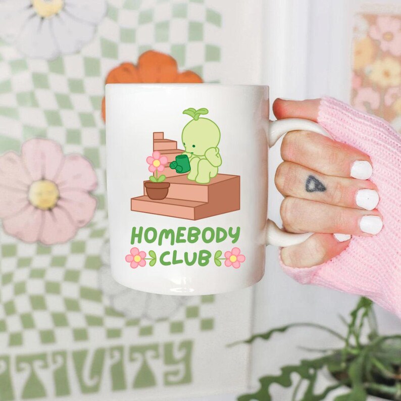 May include: White ceramic mug featuring a cartoon illustration of a green character watering a flower on a set of steps. The mug has the text "HOMEBODY CLUB" in green and pink. The mug is held by a person wearing a pink sweater.
