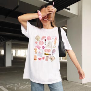May include: White t-shirt with a pink and white graphic print of various items, including a heart-shaped eye mask, a pink bunny, a pink hair clip, a pink purse, and a pink phone case.