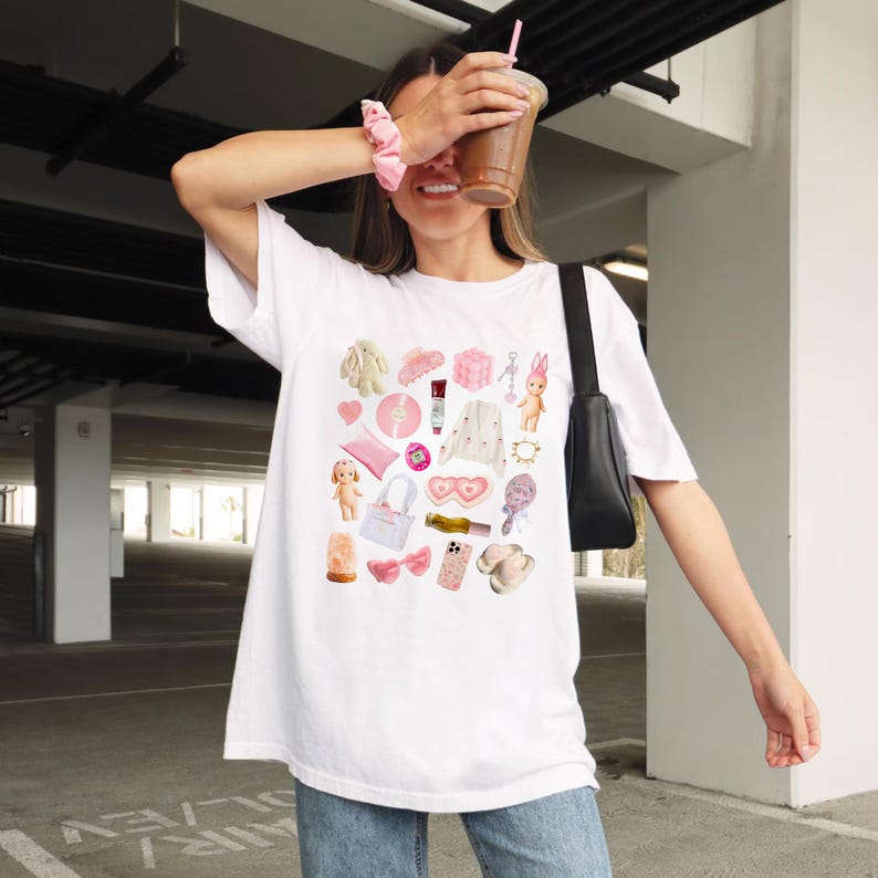 May include: White t-shirt with a pink and white graphic print of various items, including a heart-shaped eye mask, a pink bunny, a pink hair clip, a pink purse, and a pink phone case.