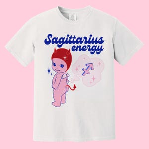 Puede incluir: Camiseta blanca con un gr&aacute;fico azul y rojo de un diablo de dibujos animados con un sombrero rojo y una burbuja de pensamiento con el s&iacute;mbolo del zod&iacute;aco Sagitario. El texto "Sagittarius energy" est&aacute; impreso en azul en la camiseta.