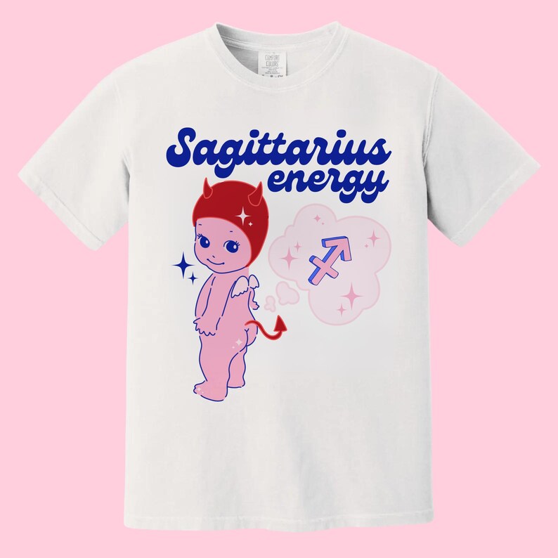 Puede incluir: Camiseta blanca con un gr&aacute;fico azul y rojo de un diablo de dibujos animados con un sombrero rojo y una burbuja de pensamiento con el s&iacute;mbolo del zod&iacute;aco Sagitario. El texto "Sagittarius energy" est&aacute; impreso en azul en la camiseta.