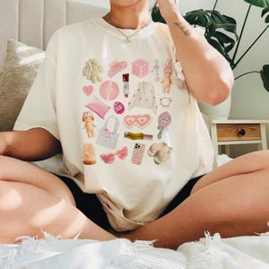 May include: A white t-shirt with a pink and white graphic print of various items, including a bunny, a hair clip, a compact mirror, a heart-shaped eye mask, a phone case, a doll, a ring, a purse, a lipstick, a nail polish bottle, a pink bow, and a pink and white cardigan.