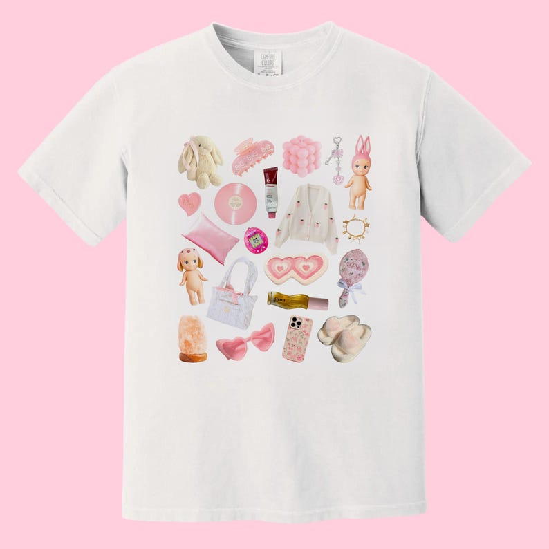 May include: A white t-shirt with a pink and white design featuring a variety of items, including a doll, a phone, a record, a heart-shaped eye mask, a bottle of perfume, a pair of slippers, and a hair clip.