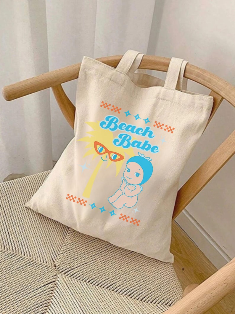 May include: Beige canvas tote bag with the words "Beach Babe" in blue and yellow. The design includes a cartoon sun wearing sunglasses and a baby in a blue bonnet. The bag has two handles and is sitting on a wooden chair.